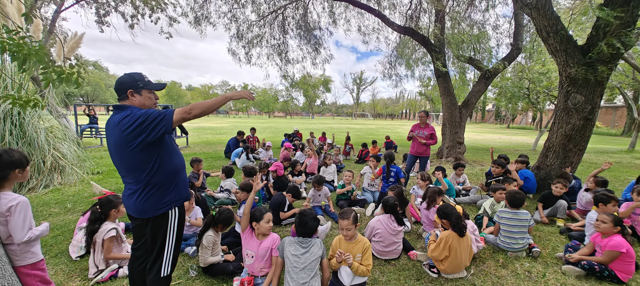 Grupo de niños sentados en el pasto bajo un árbol, atendiendo a su maestro