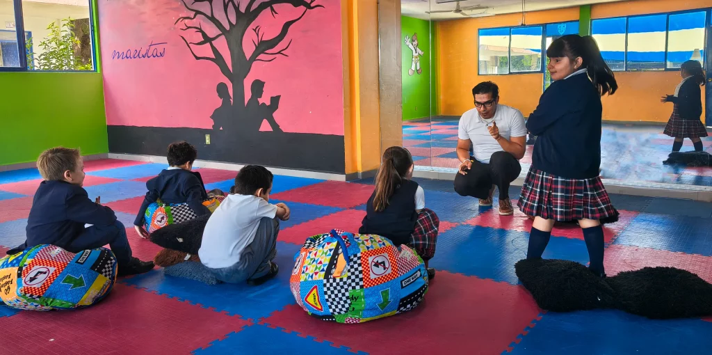 Alumnos sentados sobre tapetes de colores en el salón de Interioridad, con su maestro